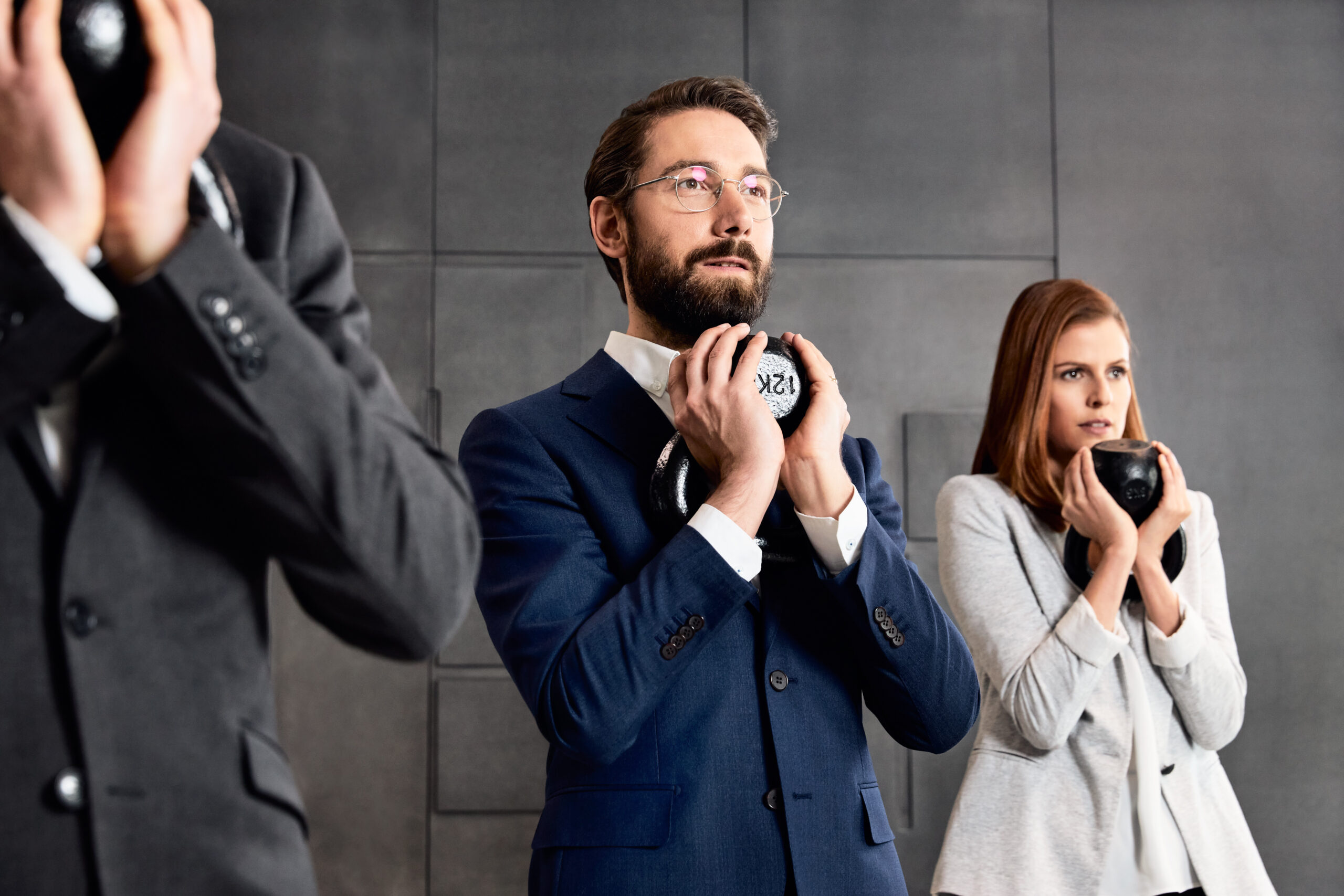 Drei Menschen stehen in Business-Kleidung im Fitness-Studio und haben Hanteln in der Hand.
