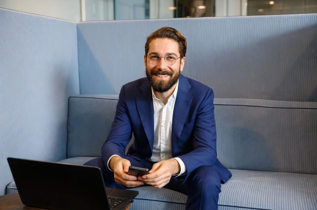 Chris Schiebel sitzt lächelnd mit Smartphone und Laptop auf einem blauen Sofa